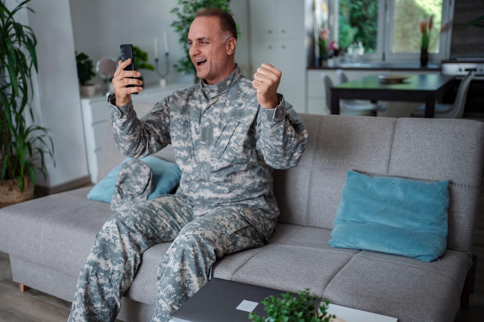 Soldier celebrating good news on smartphone while sitting on sofa at home